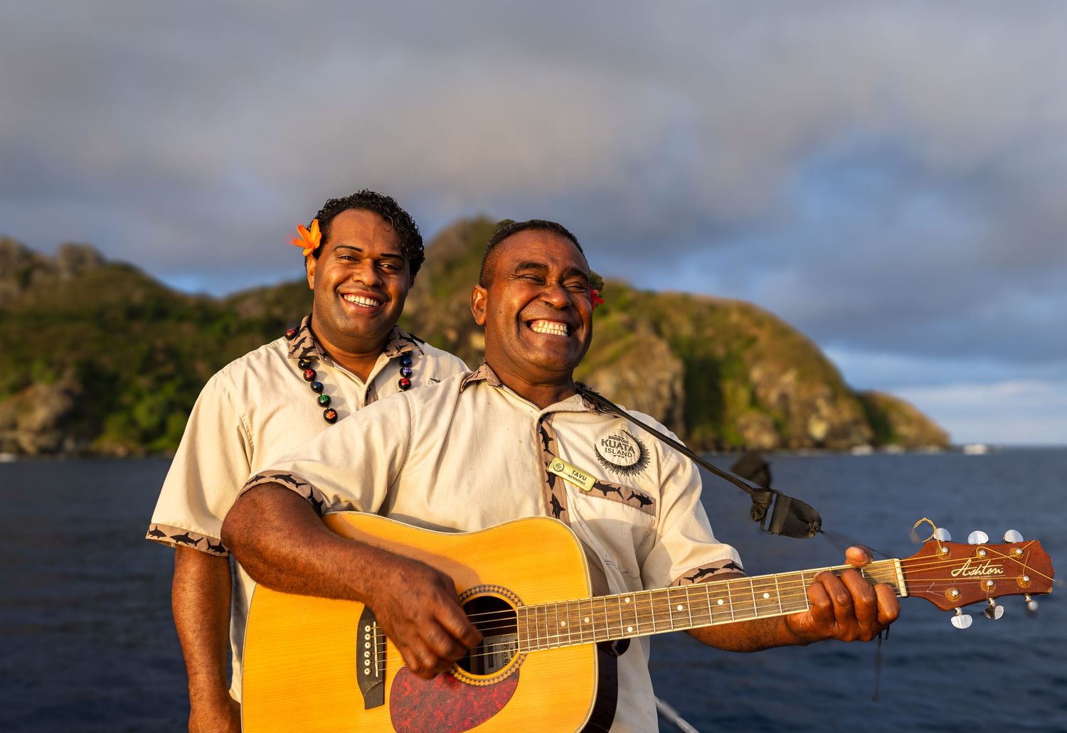 Two musicians perform together with a guitar against a tropical coastal backdrop at sunset.