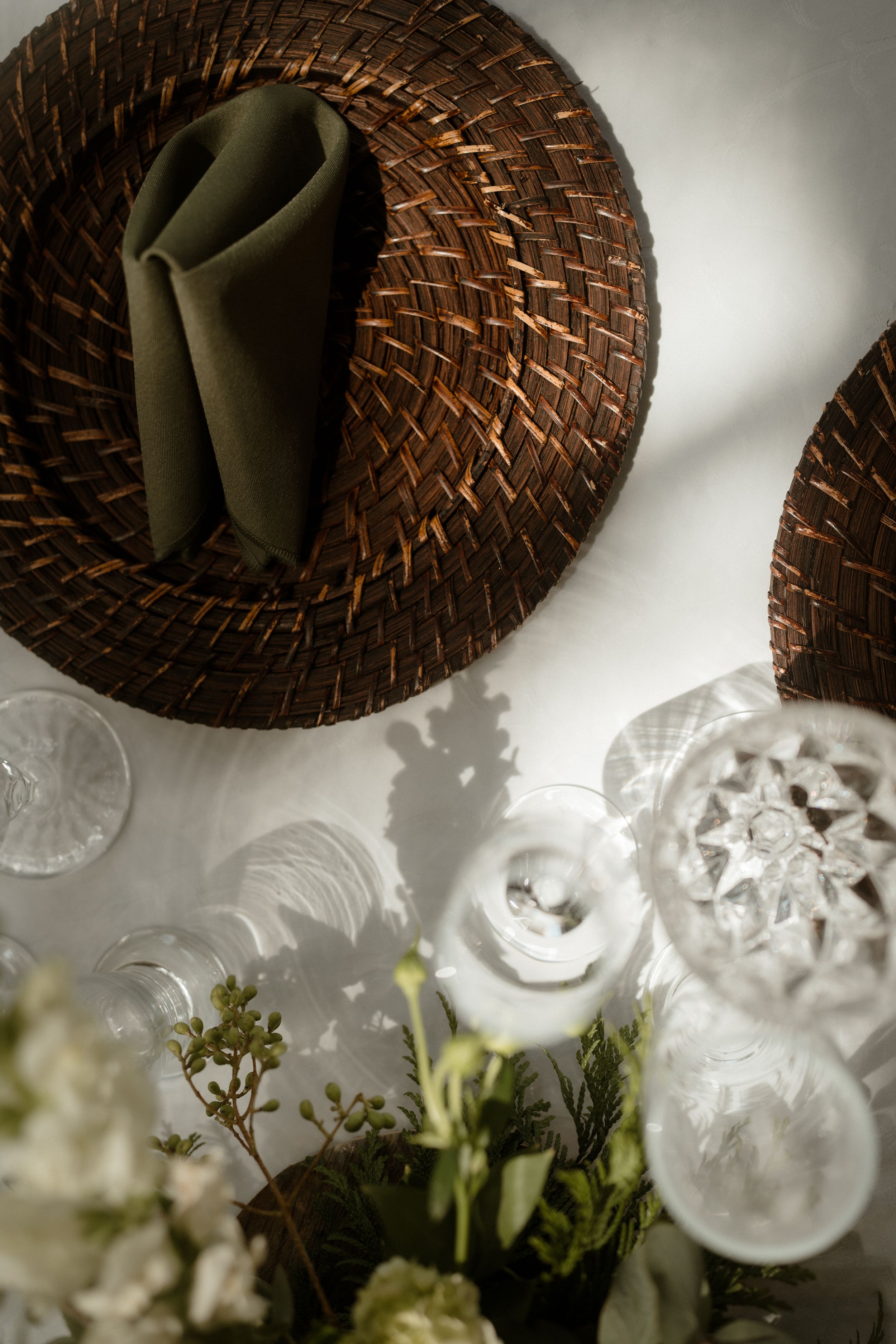 Rustic woven charger plates and olive napkins on white tablecloth with floral accents.