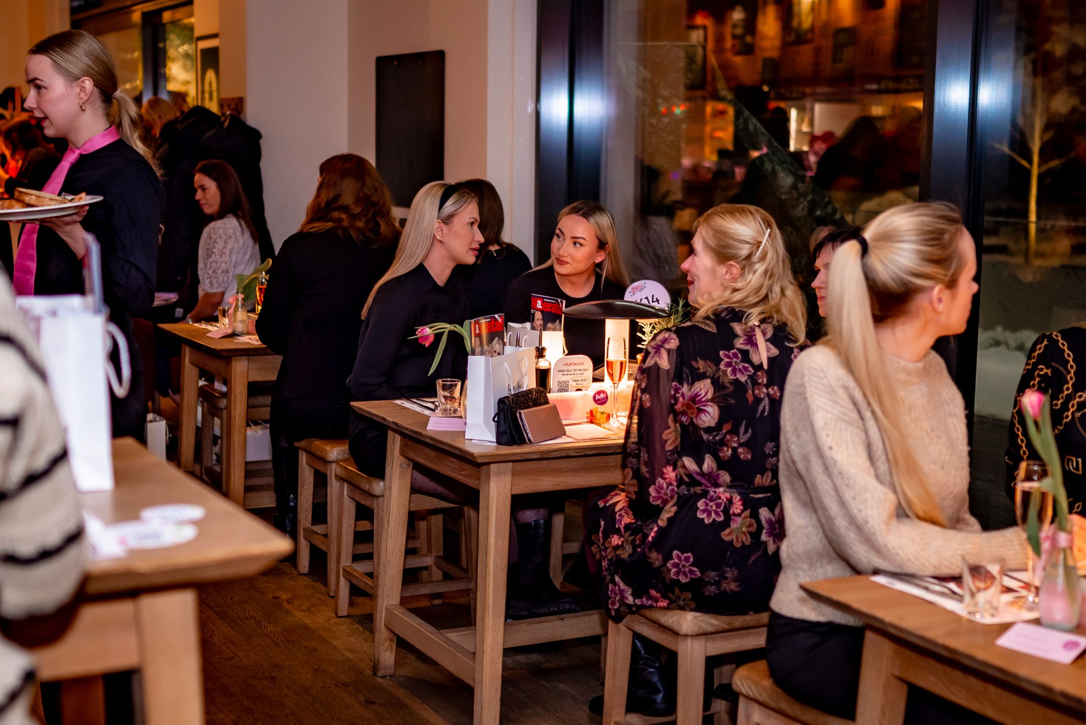 Restaurant guests enjoying dinner and drinks at wooden tables in a warm, dimly lit dining atmosphere.