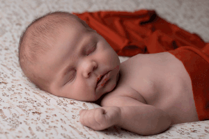 Newborn girl draped in orange fabric, sleeping on a floral background during a professional newborn session in Ceredigion