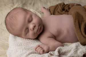 Newborn pouting while wrapped in a brown cheesecloth wrap, while sleeping in a wooden prop during a newborn session in Ceredigion