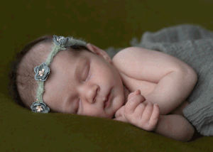 Newborn girl wearing a green floral headband and wrapped in a green cheesecloth wrap, sleeping on a dark green background in Ceredigion