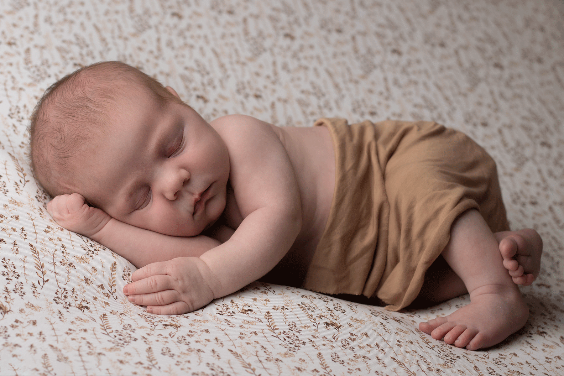 Newborn wrapped in brown wrap, sleeping peacefully on a floral background during a professional newborn photo session in Ceredigion