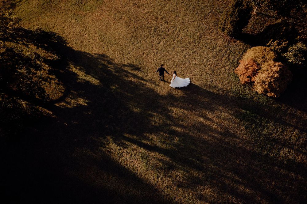 Aerial view of bride and groom walking across a field, white wedding dress trailing behind.