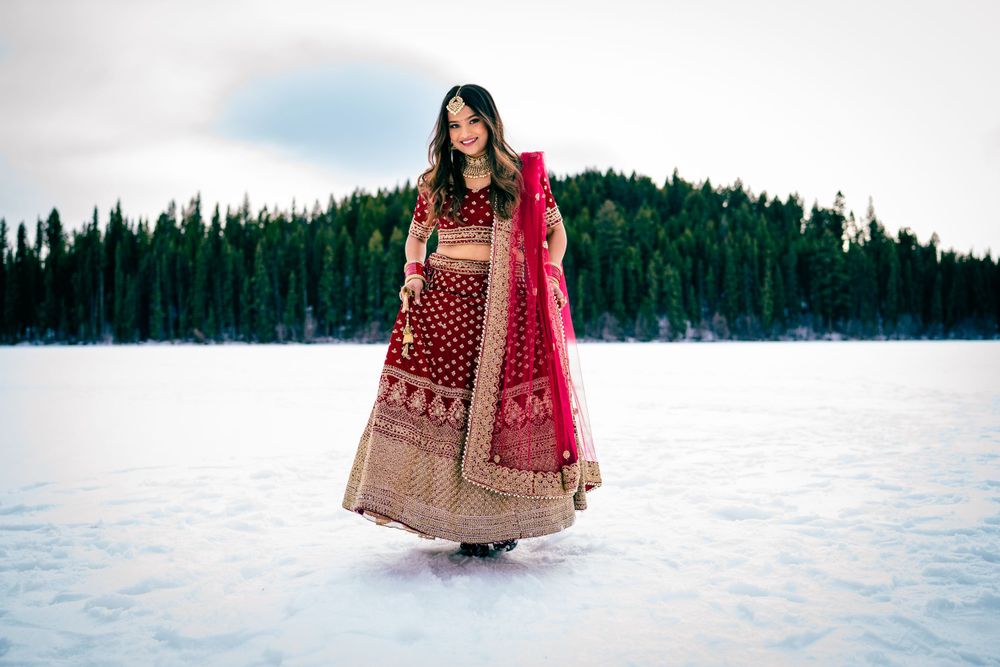 Traditional Indian bride in red lehenga standing in snowy landscape with evergreen trees.