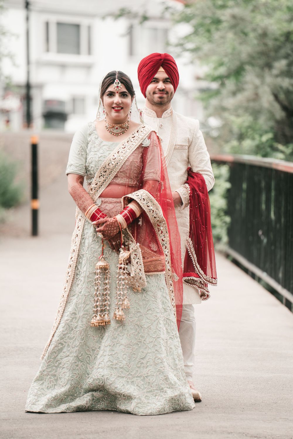 Couple in traditional Sikh wedding attire posing on urban bridge.