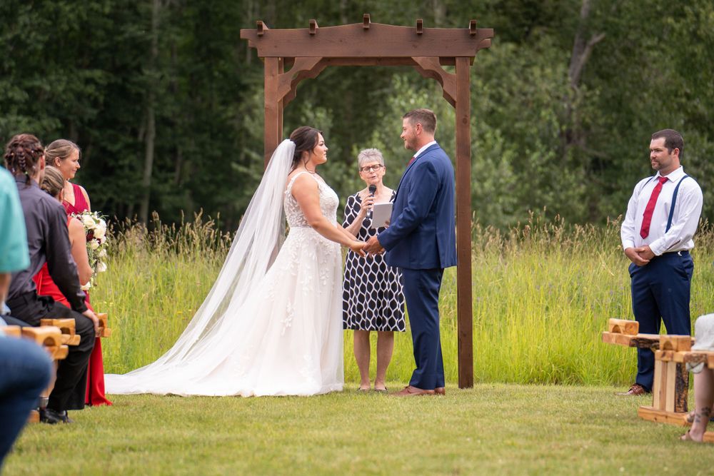 Outdoor wedding ceremony under wooden arch with guests seated on benches.