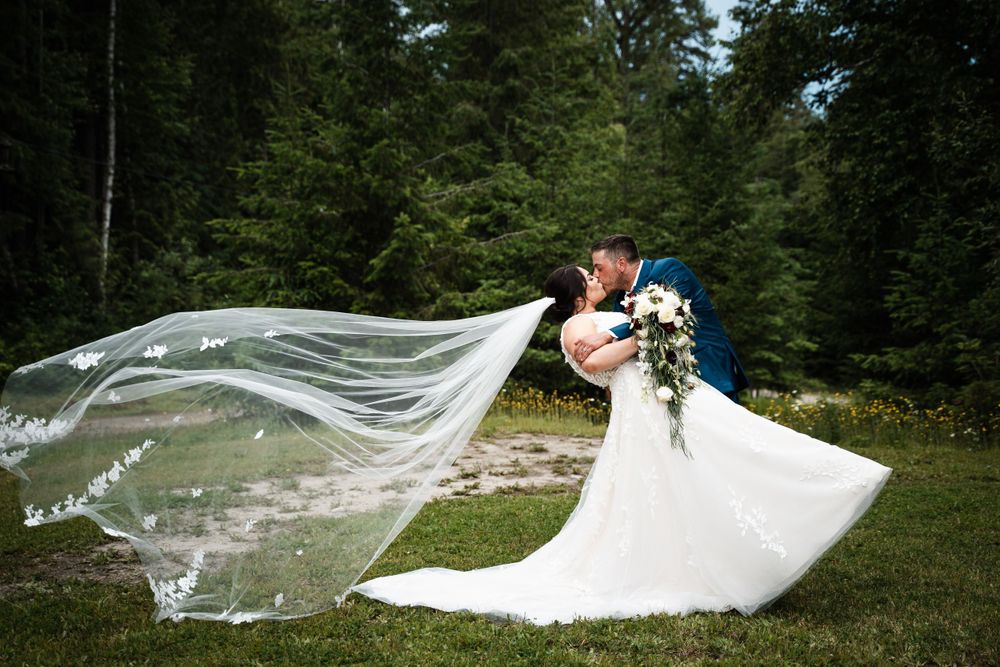 A wedding couple embraces in a forest setting as a long veil billows dramatically in the breeze.