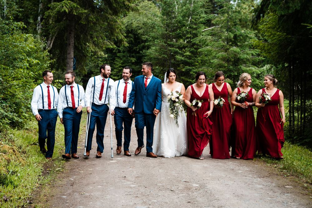 Wedding party poses on forest path with bridesmaids in red dresses and groomsmen in suspenders.