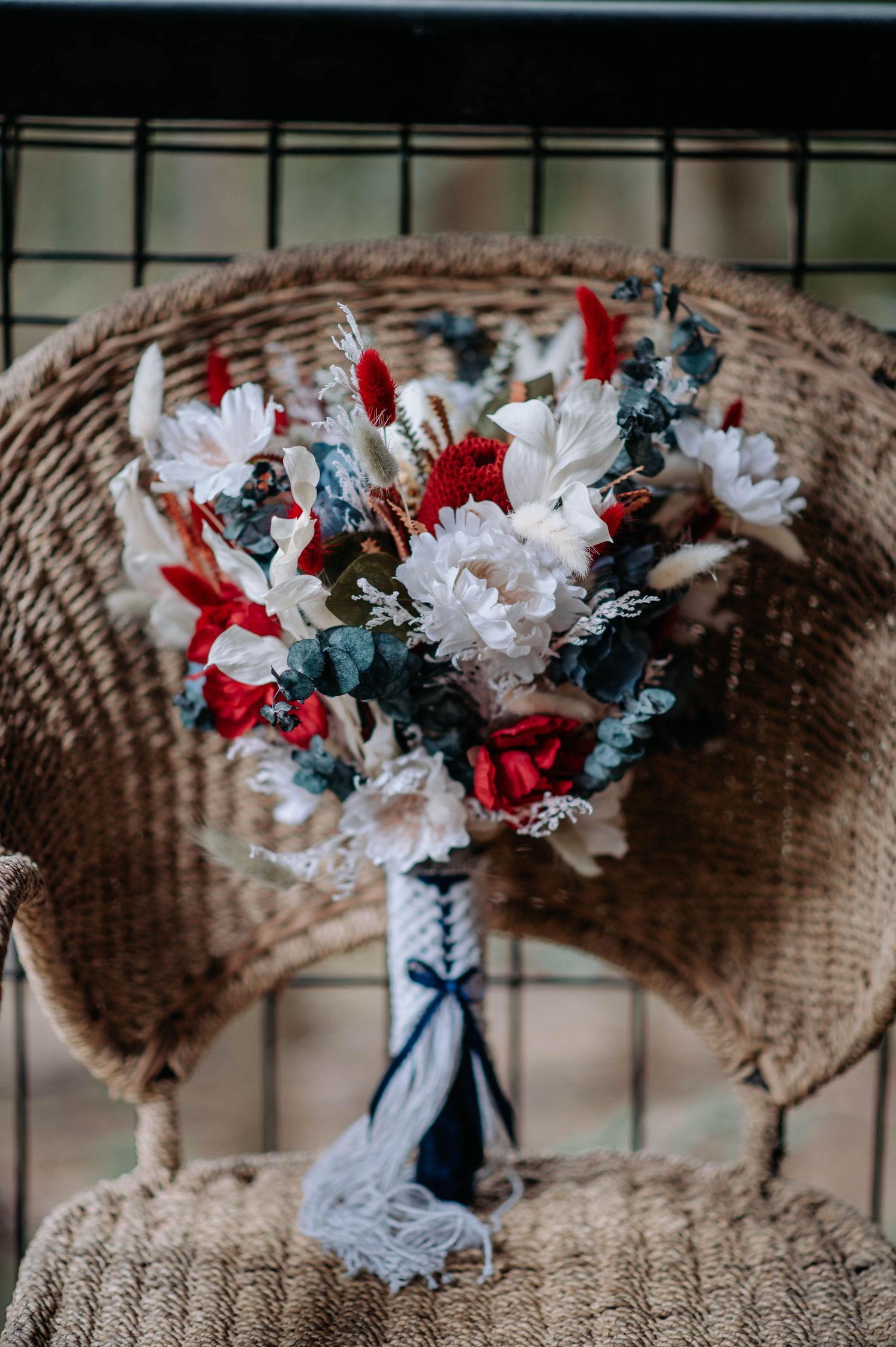A rustic wedding bouquet with red, white and blue flowers displayed on a woven chair.