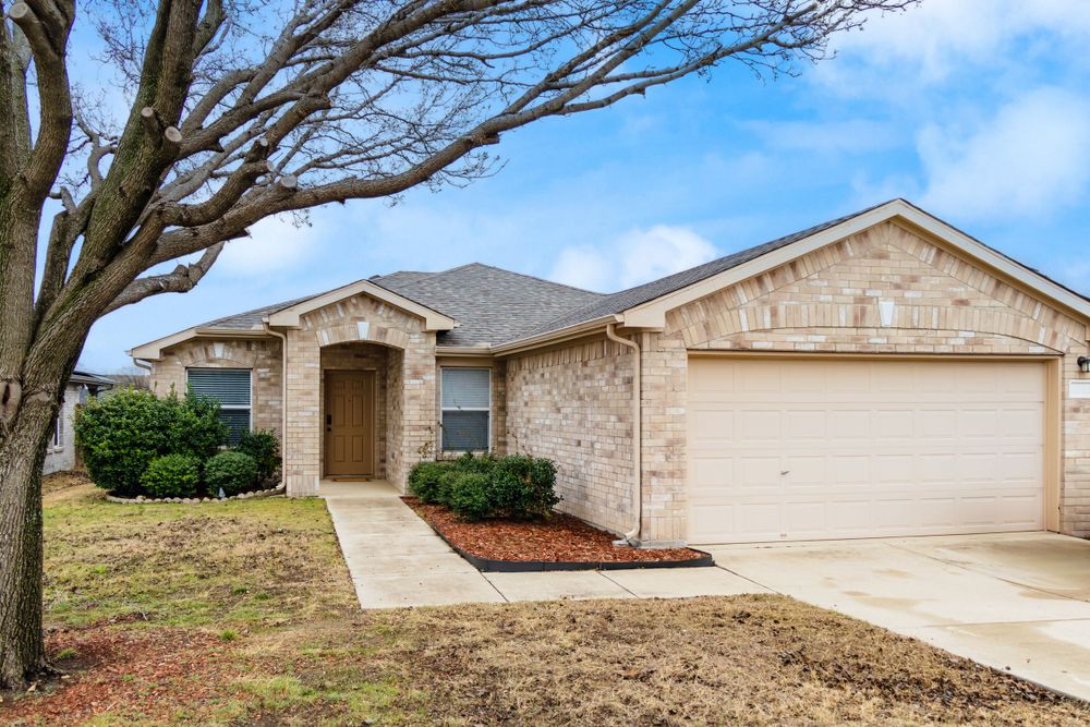 Beige brick ranch house with attached garage and landscaping under cloudy sky.