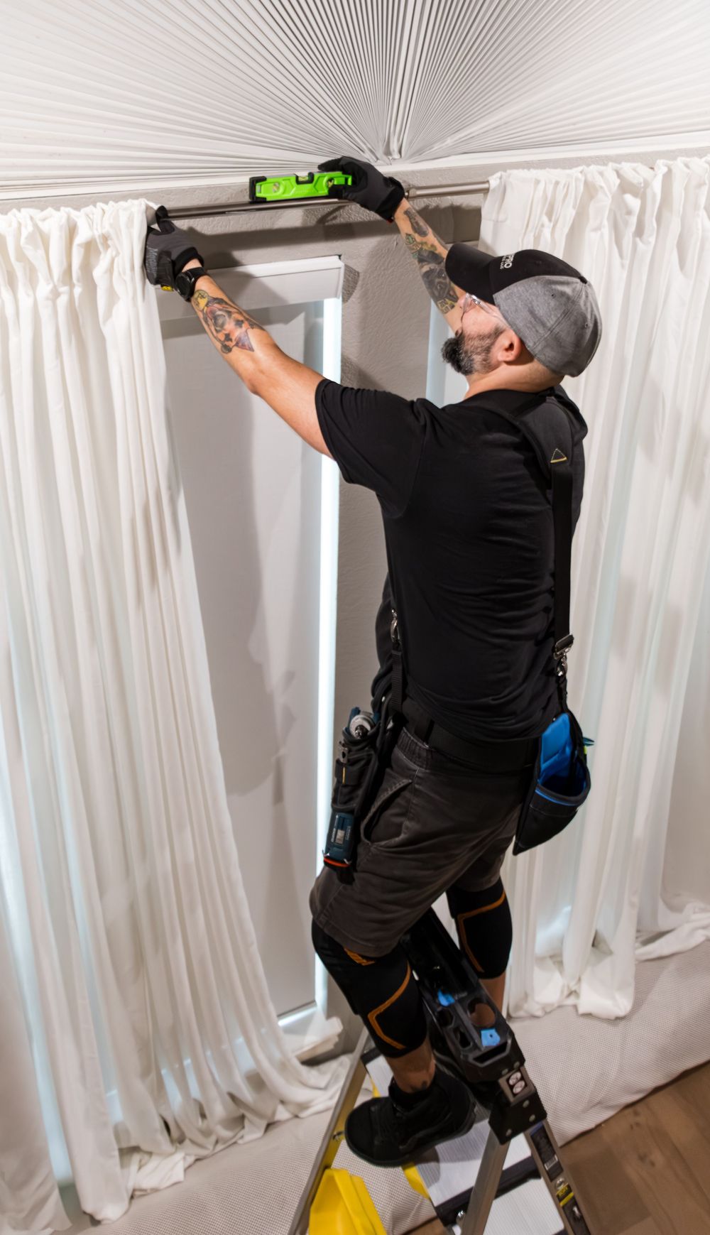 Tattooed installer on ladder using level to mount curtain rod between white drapes in modern interior.