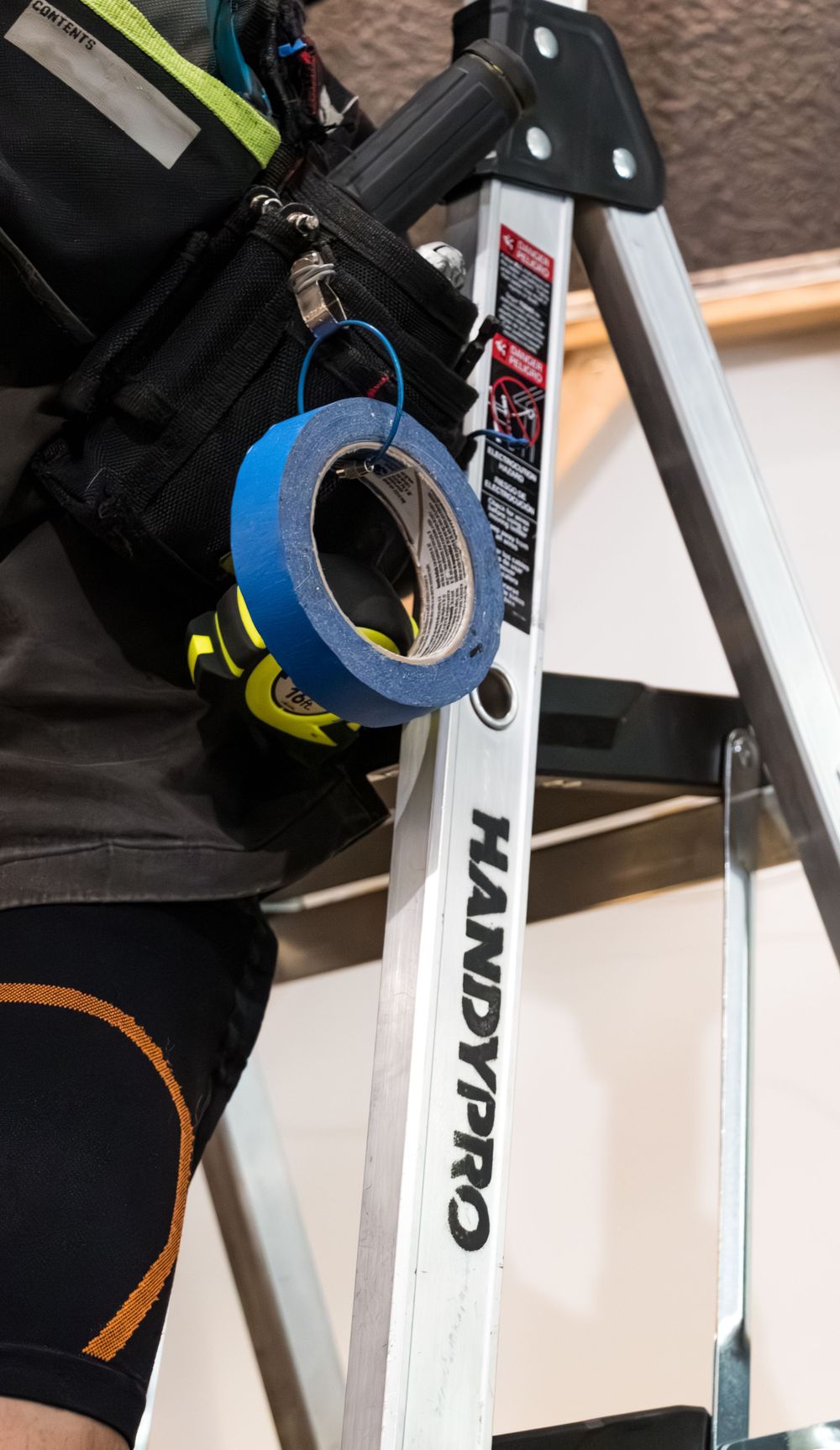 Worker on a HandyPro ladder with blue painter's tape and tools on a tool belt during a home improvement project.