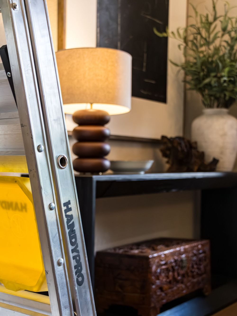 A HANDYPRO ladder leans against a wall near a wooden table lamp, decorative box, and plant on a dark shelf.
