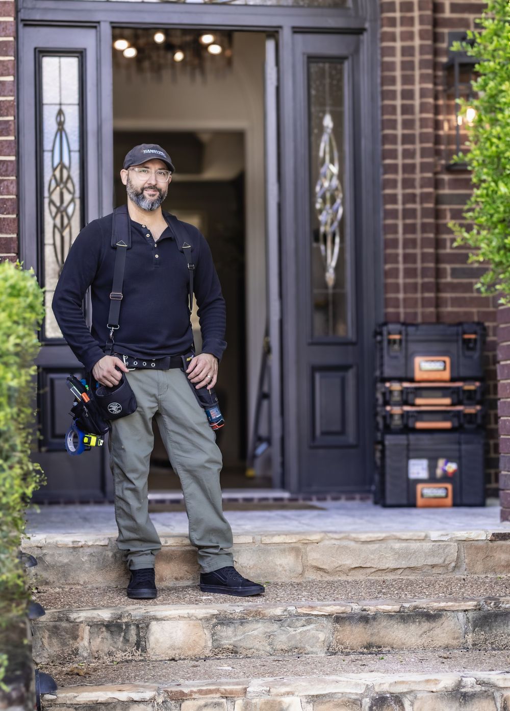 A smiling male contractor stands on stone steps outside a brick home, wearing a tool belt and holding equipment.