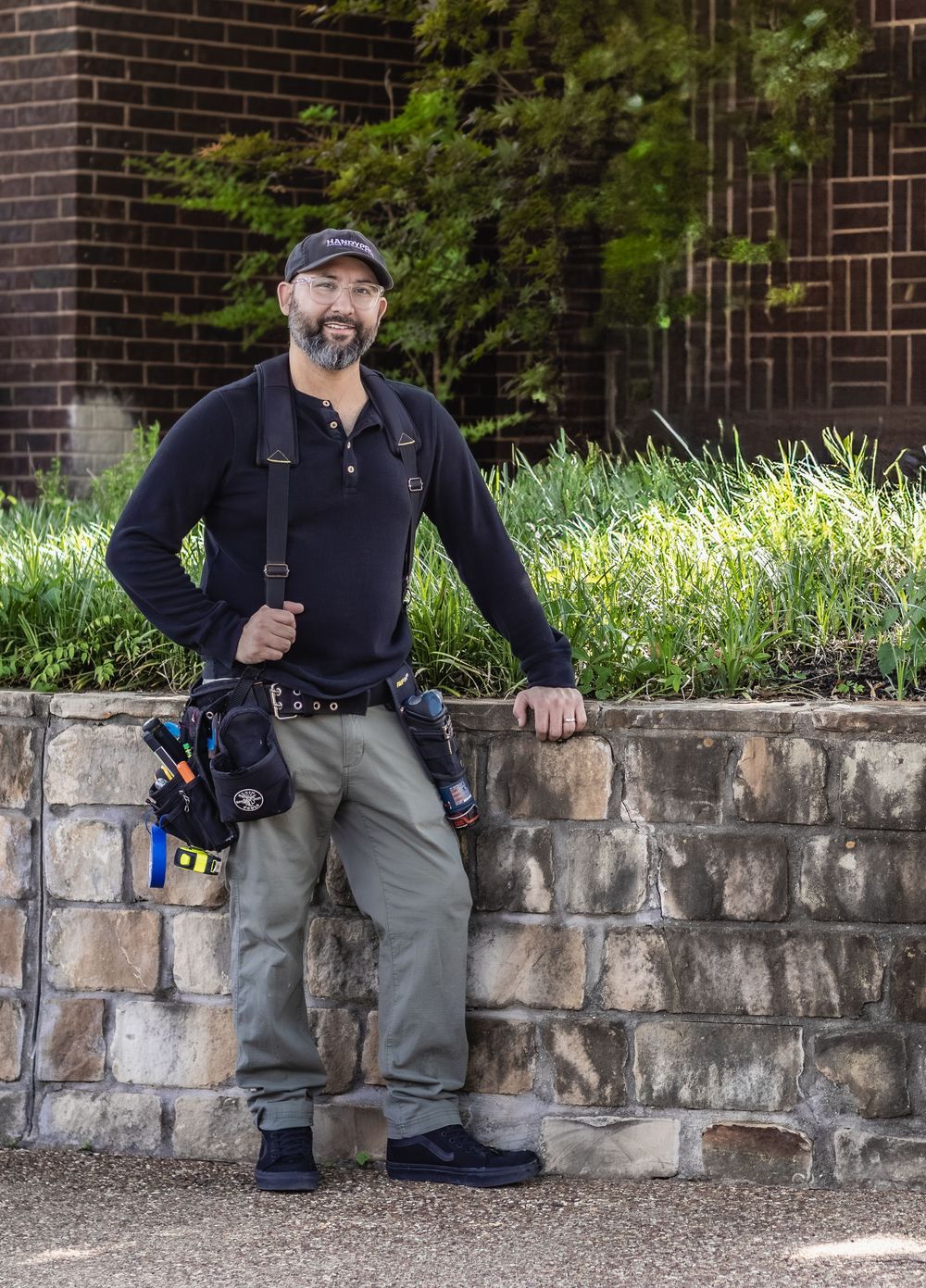 A smiling male tradesman in work gear and tool belt leans against a stone wall outdoors near green shrubs.
