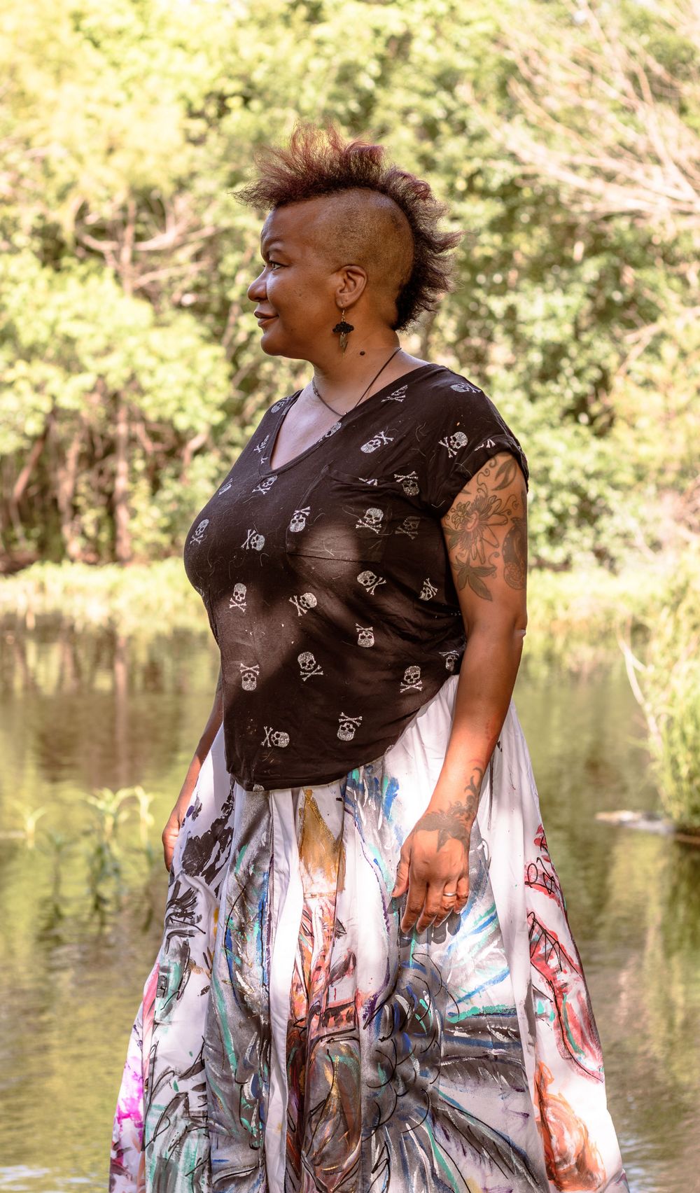 A Black woman with a mohawk hairstyle and arm tattoos poses outdoors wearing a skull-print top and colorful skirt.