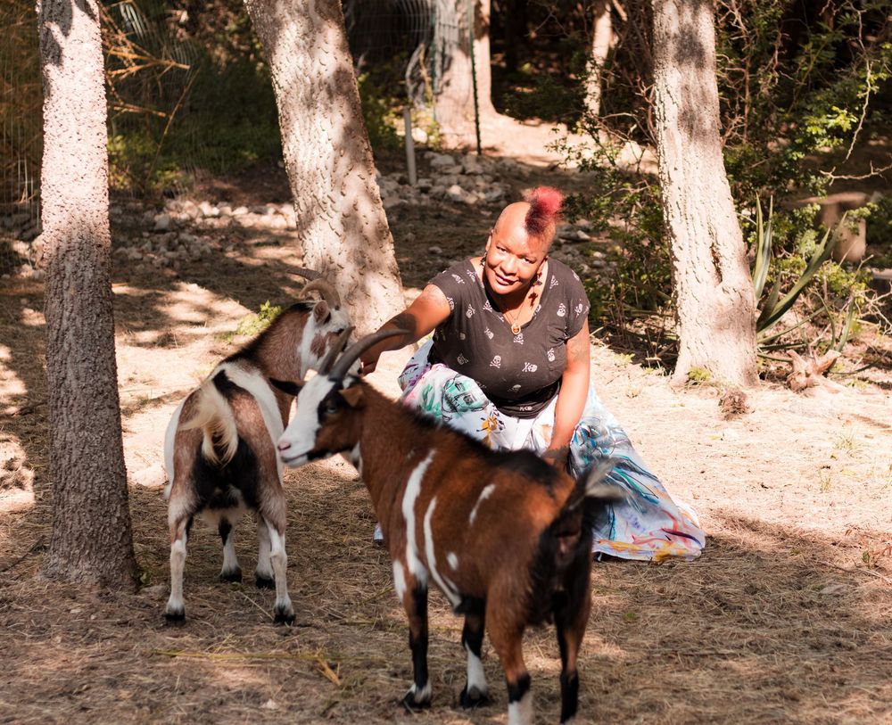 Woman with red hair petting two brown and white goats in a wooded outdoor setting.