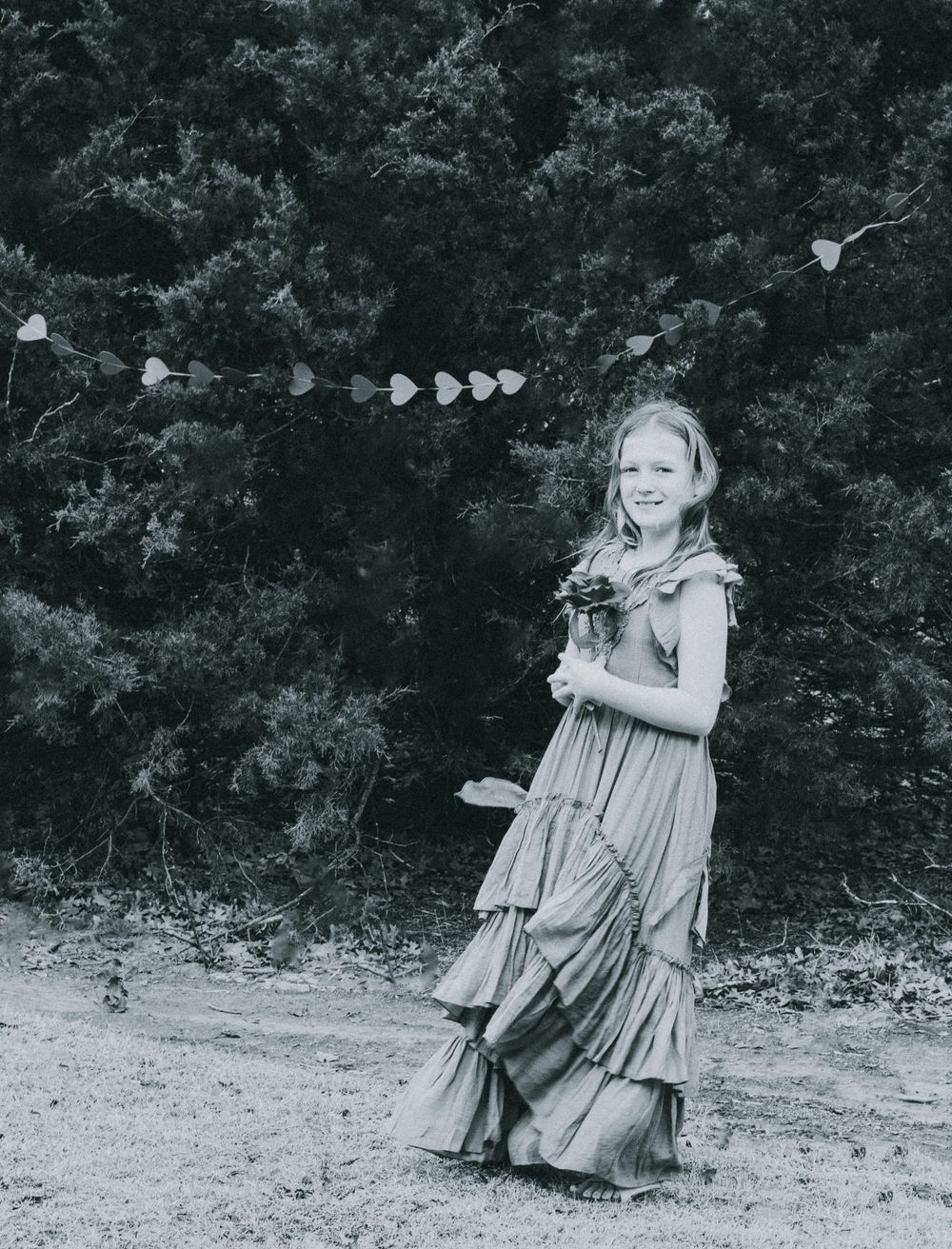 A black and white photo of a person in a vintage tiered dress standing outdoors with heart decorations.