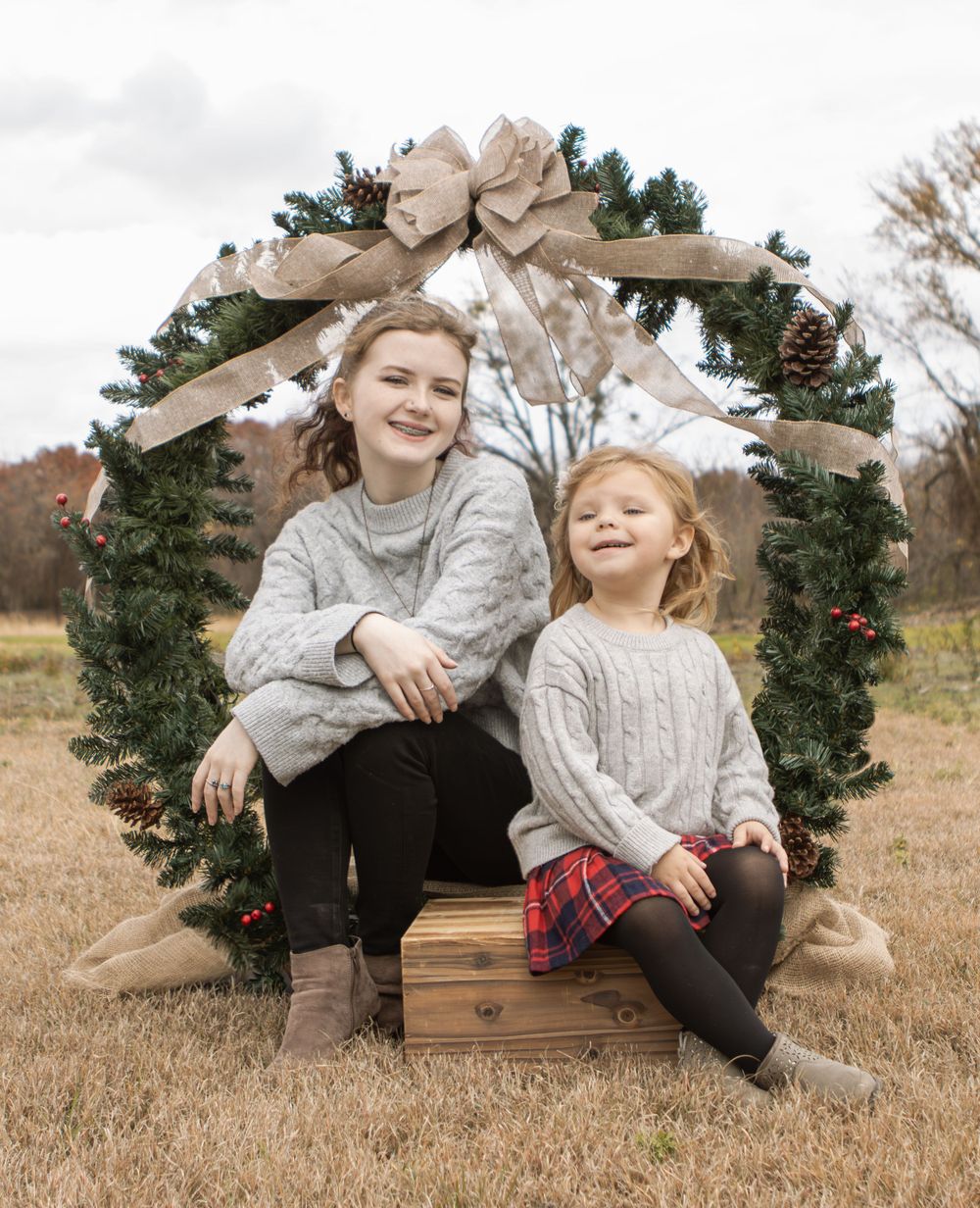 Two young children sit together on a wooden crate with a holiday wreath backdrop.