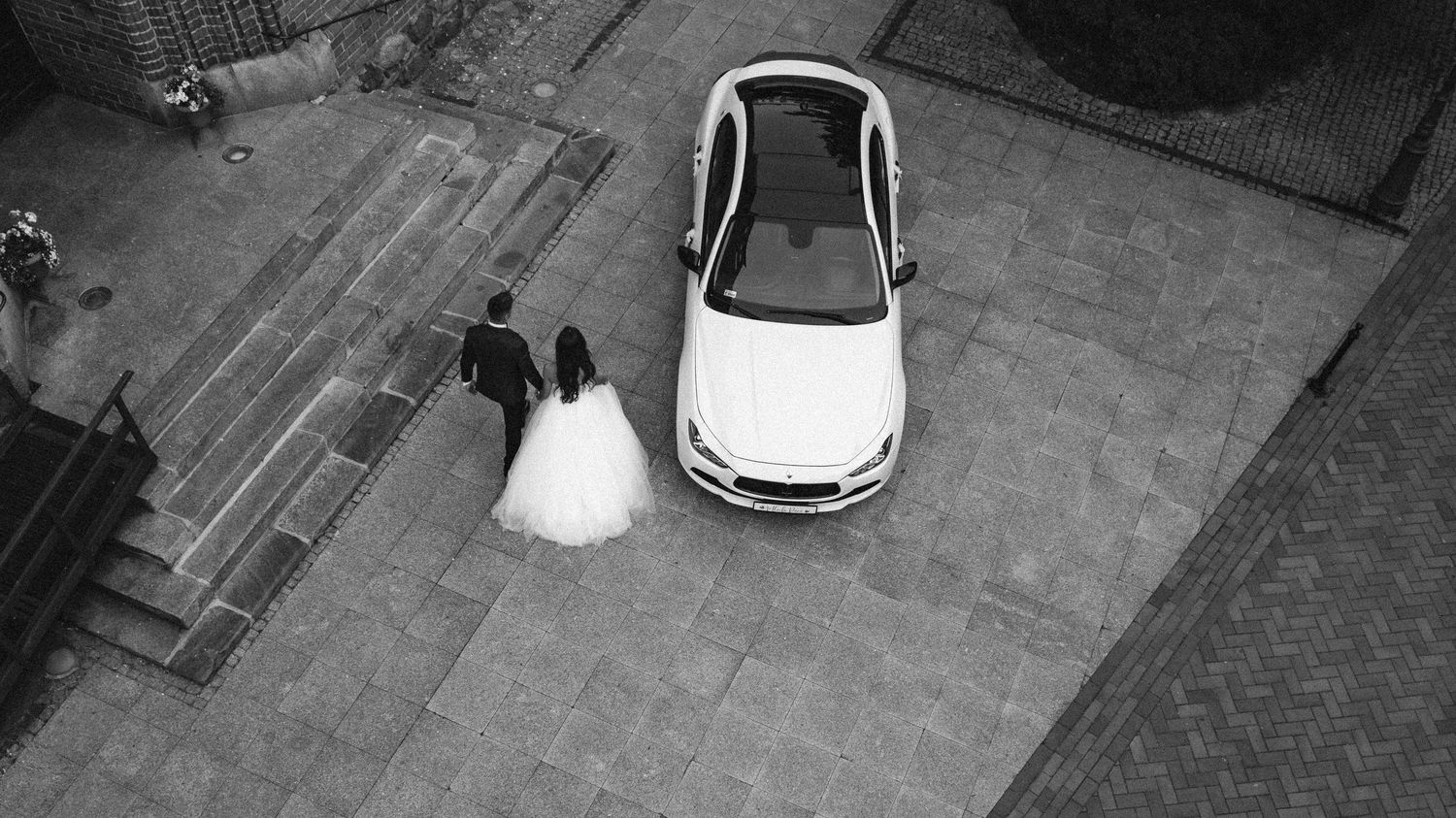 Aerial view of newlyweds walking next to a white car on paved stone ground.