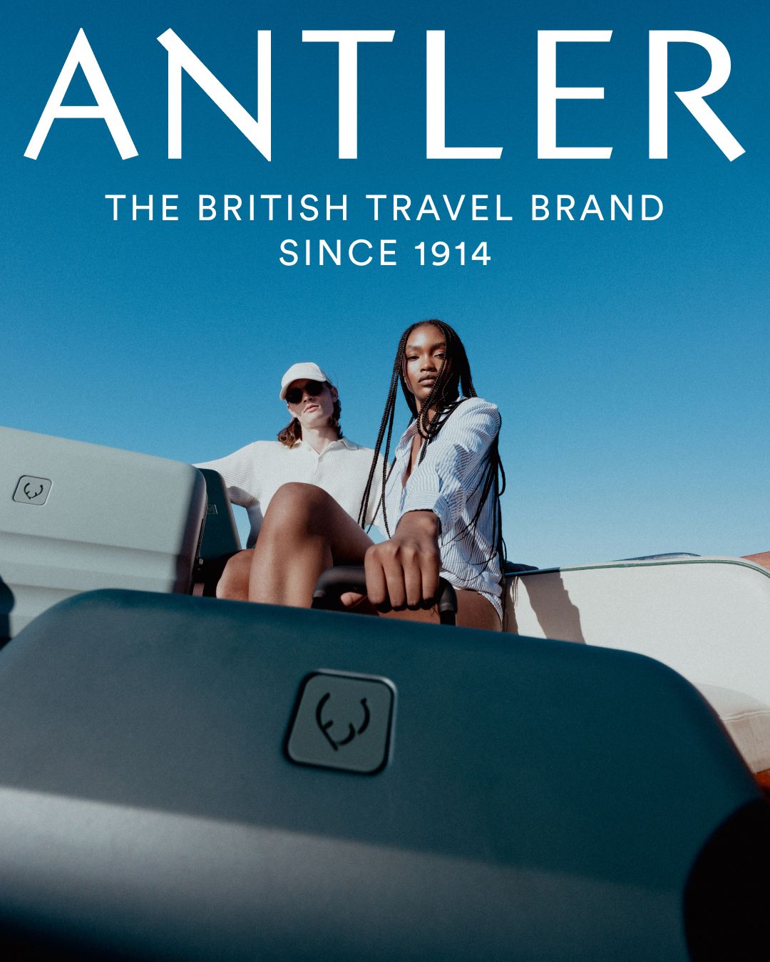 Antler brand advertising featuring two models sitting on blue luggage against desert background.