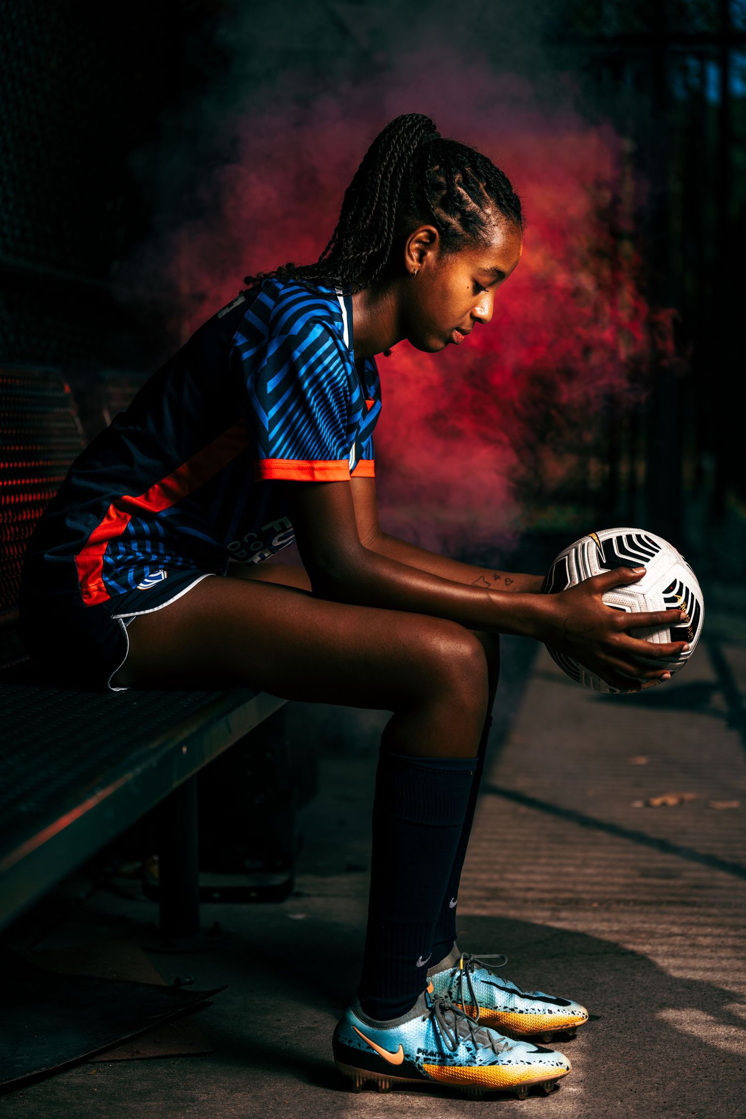 A soccer player from Reign Academy sits on a bench holding a soccer ball against a backdrop of red smoke.