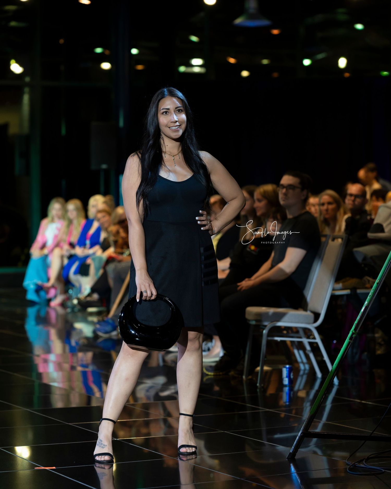 Model walks the runway in a black cocktail dress and heels during an evening fashion show.
