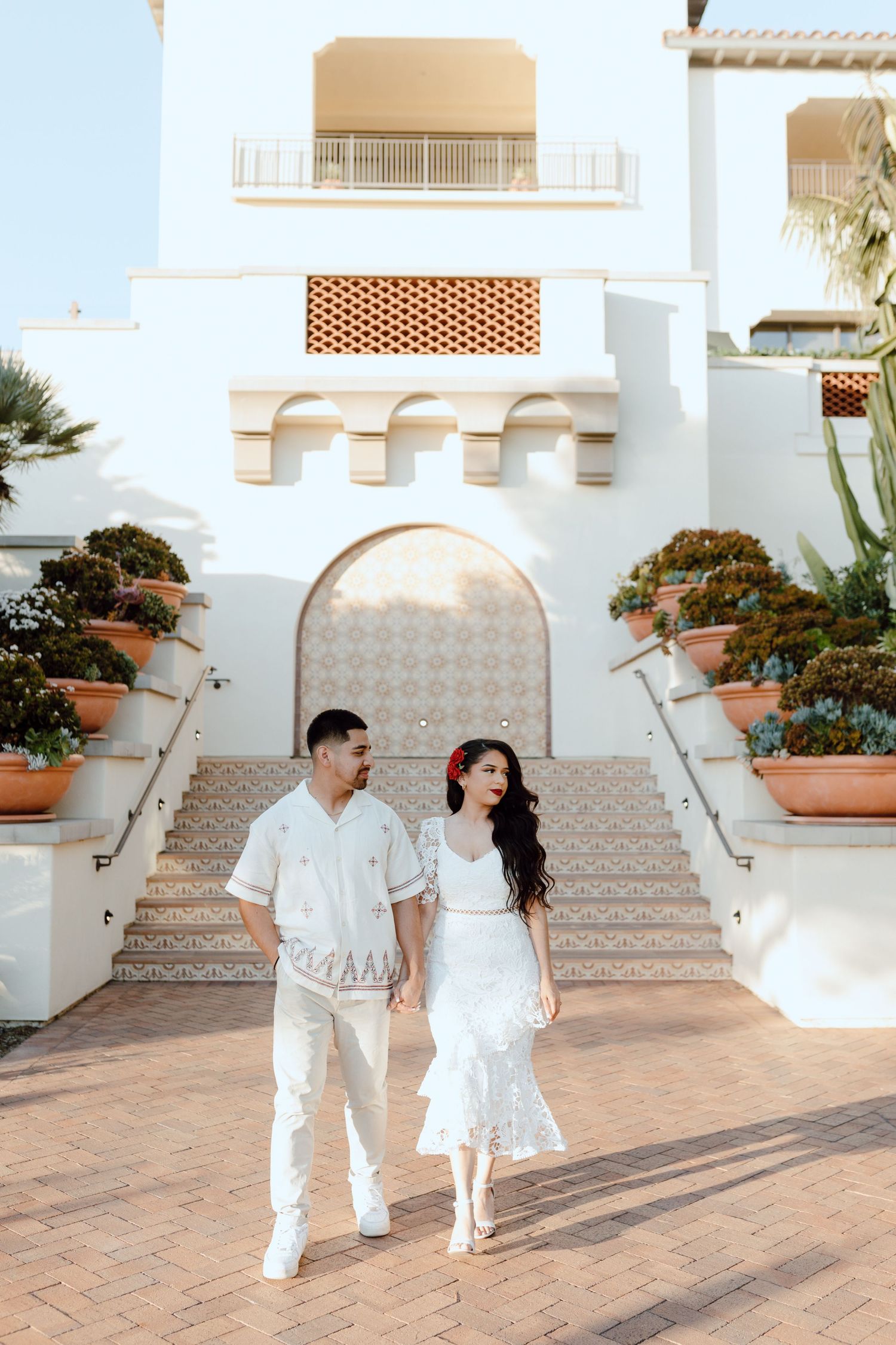 Couple in white outfits walking along Mediterranean-style steps with terracotta planters at a luxury resort.