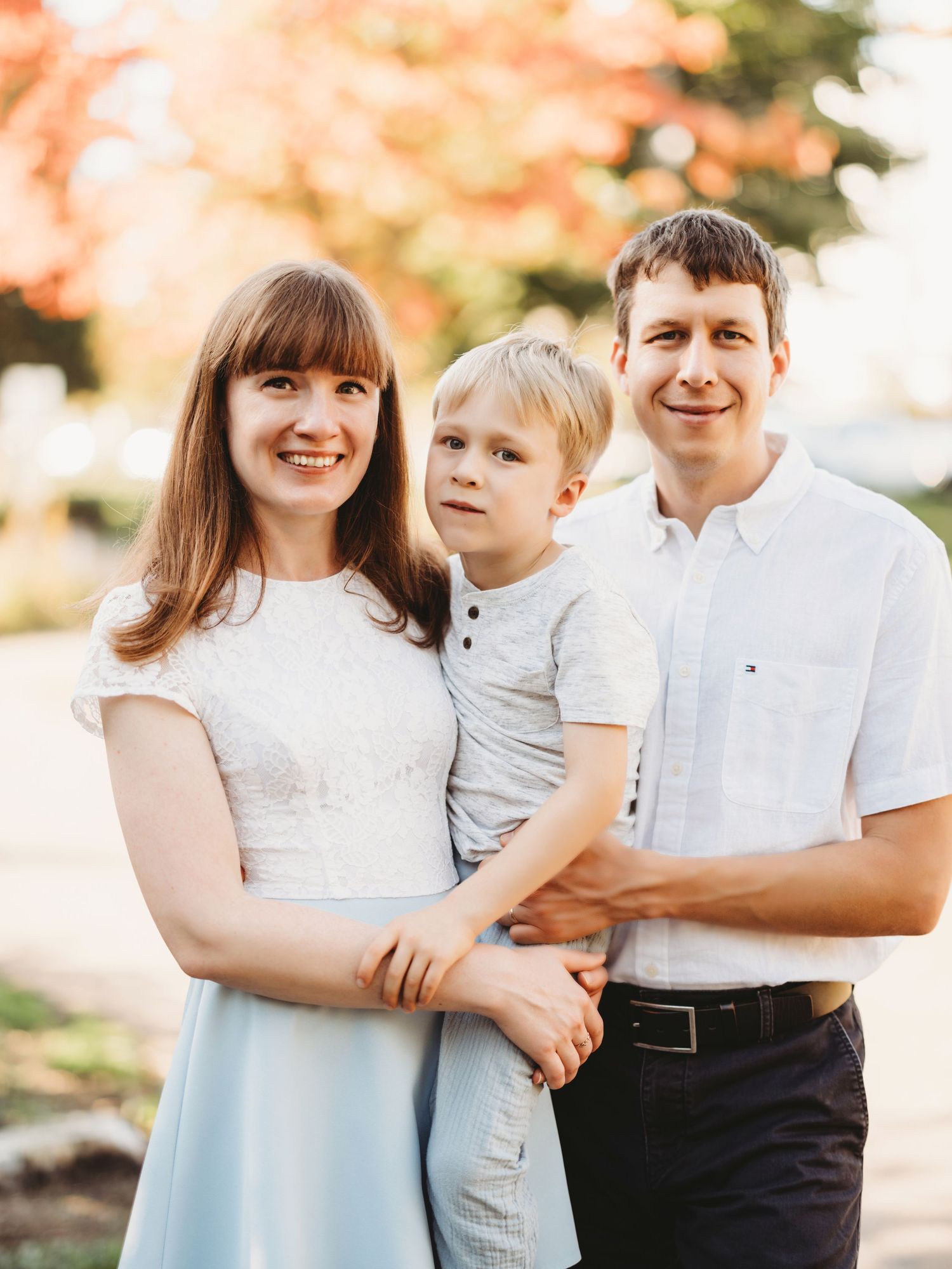 A family portrait outdoors with autumn trees and warm sunlight.