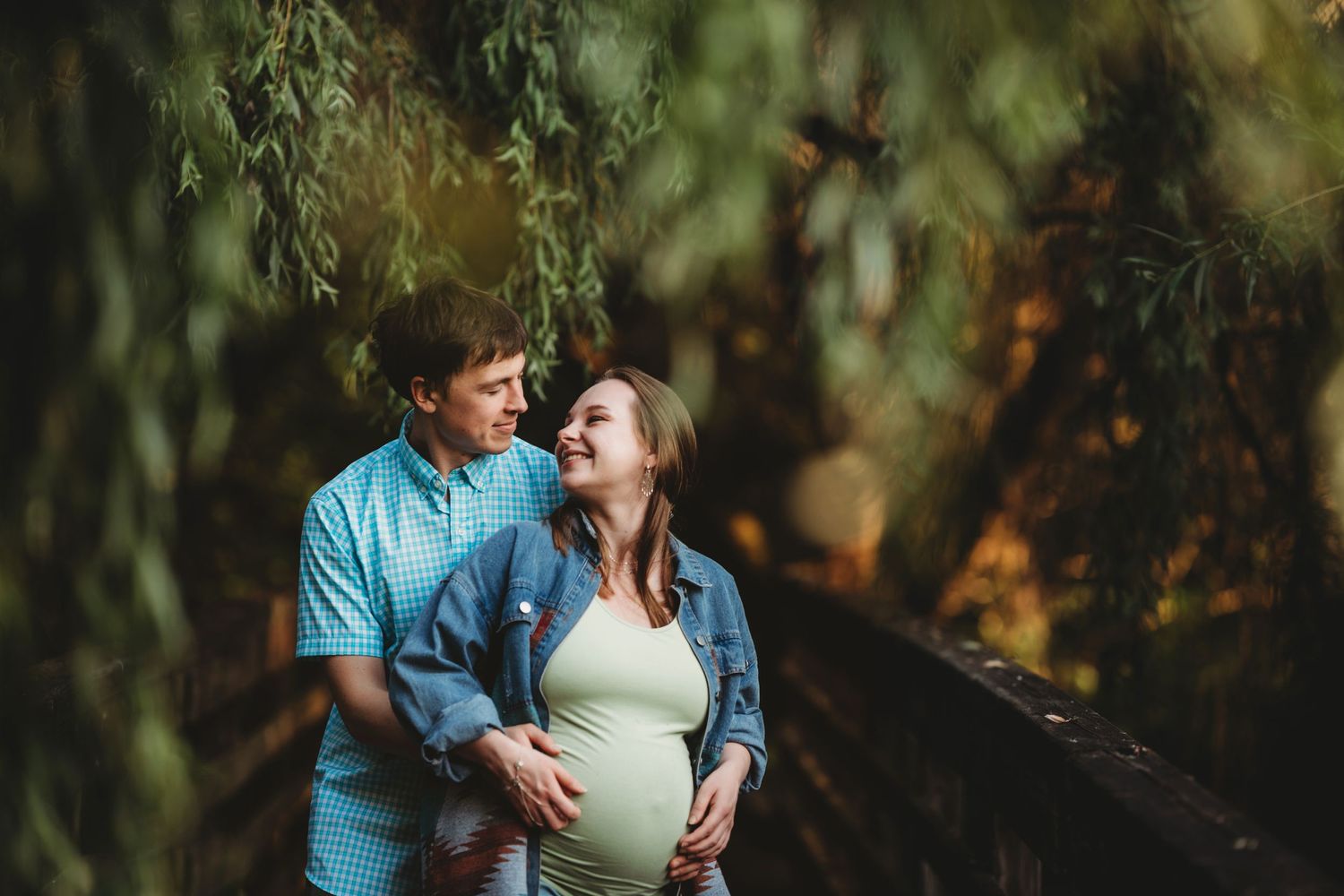 Expecting couple sharing an intimate moment under hanging willow branches in soft lighting.