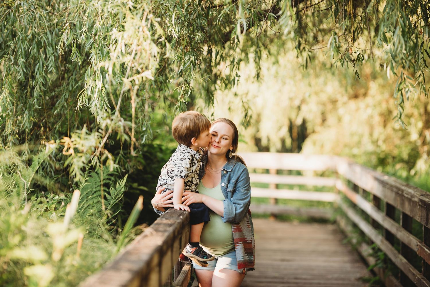 A loving embrace shared on a wooden boardwalk surrounded by lush greenery on a sunny day.