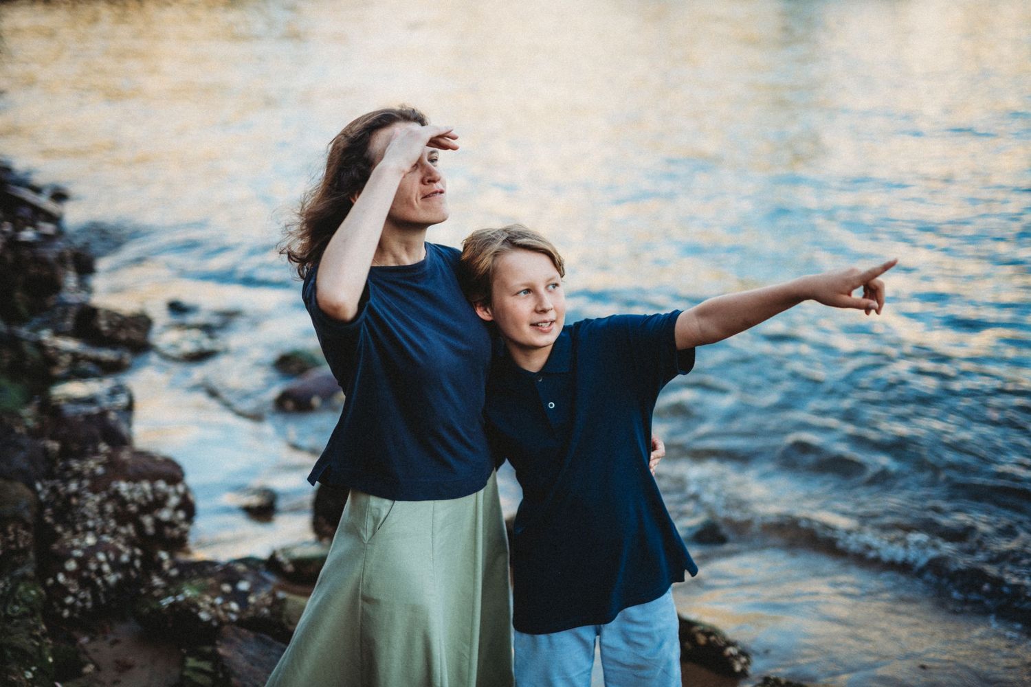 Two people pointing and enjoying a scenic ocean view from rocky coastline at sunset.