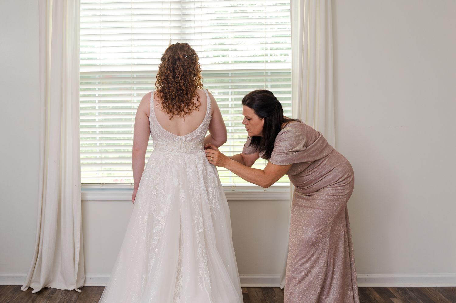 A wedding dress being fitted and adjusted in a bright room with white curtains.