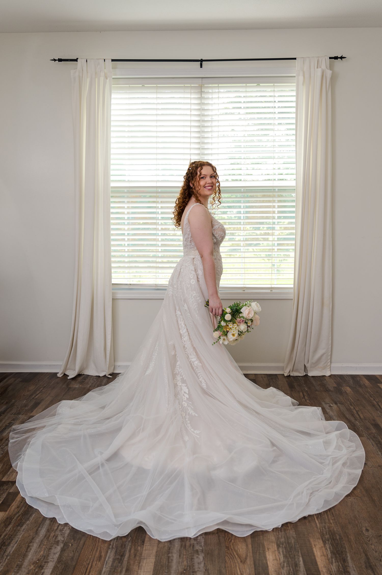 An elegant wedding gown with a flowing train photographed against white curtains in natural light.