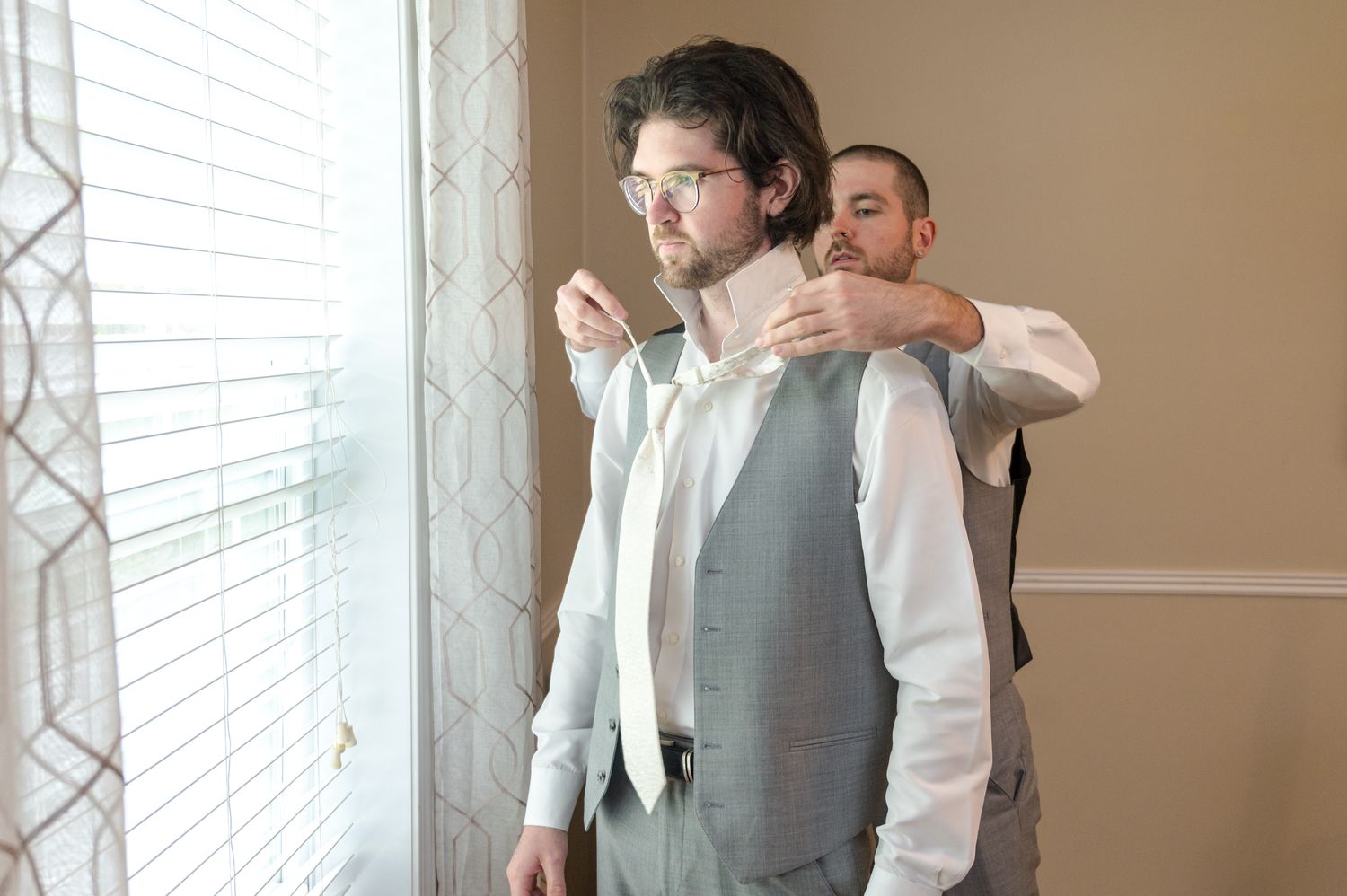 Two people helping with formal attire preparations by a window.