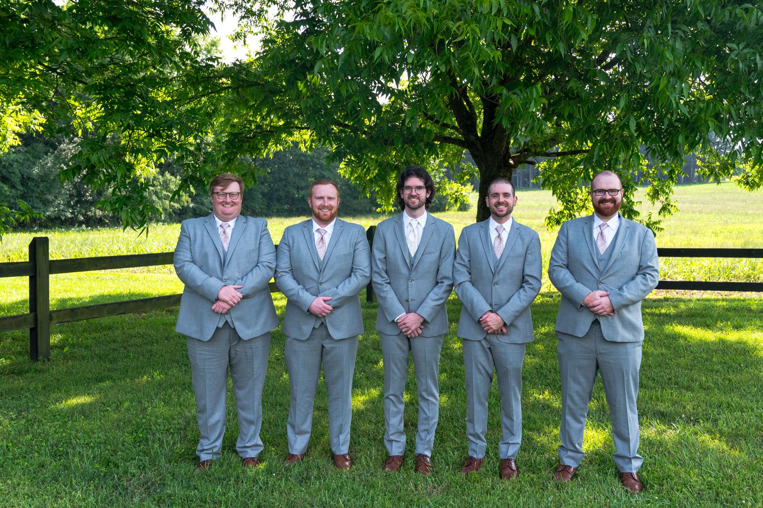 Groomsmen in matching gray suits pose together on a sunny lawn with wooden fence backdrop.