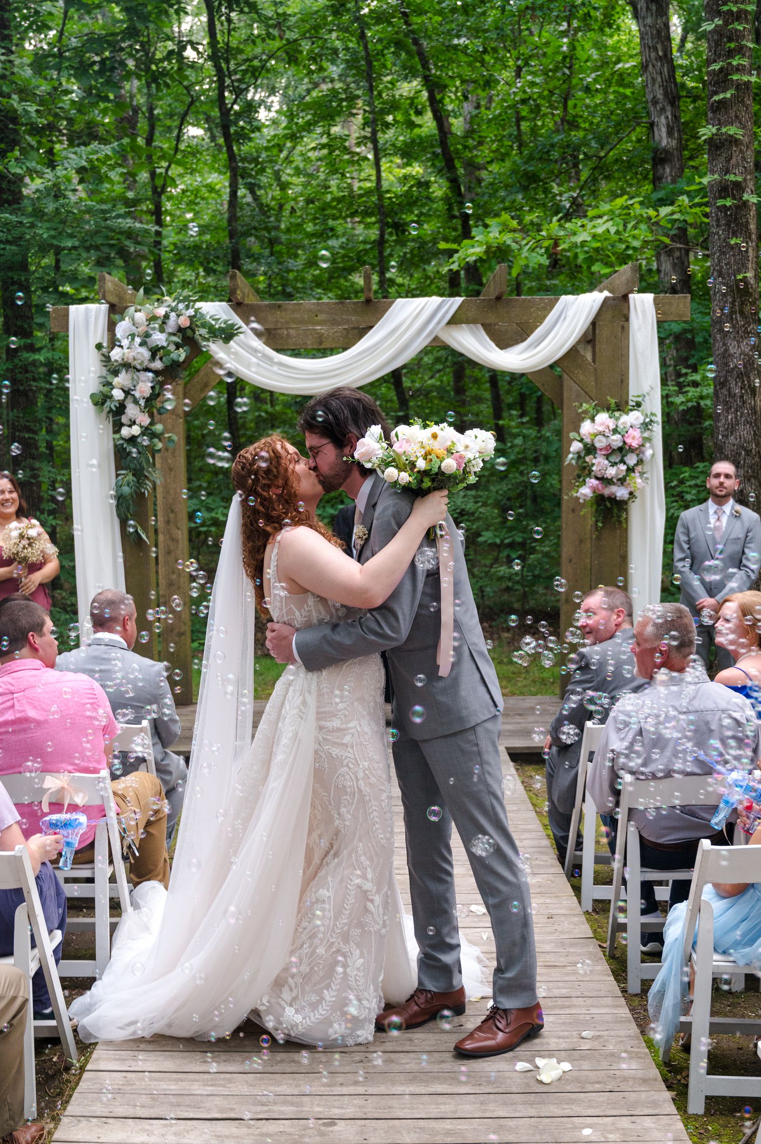 Wedding ceremony with decorated wooden arch and seated guests in a forest setting.
