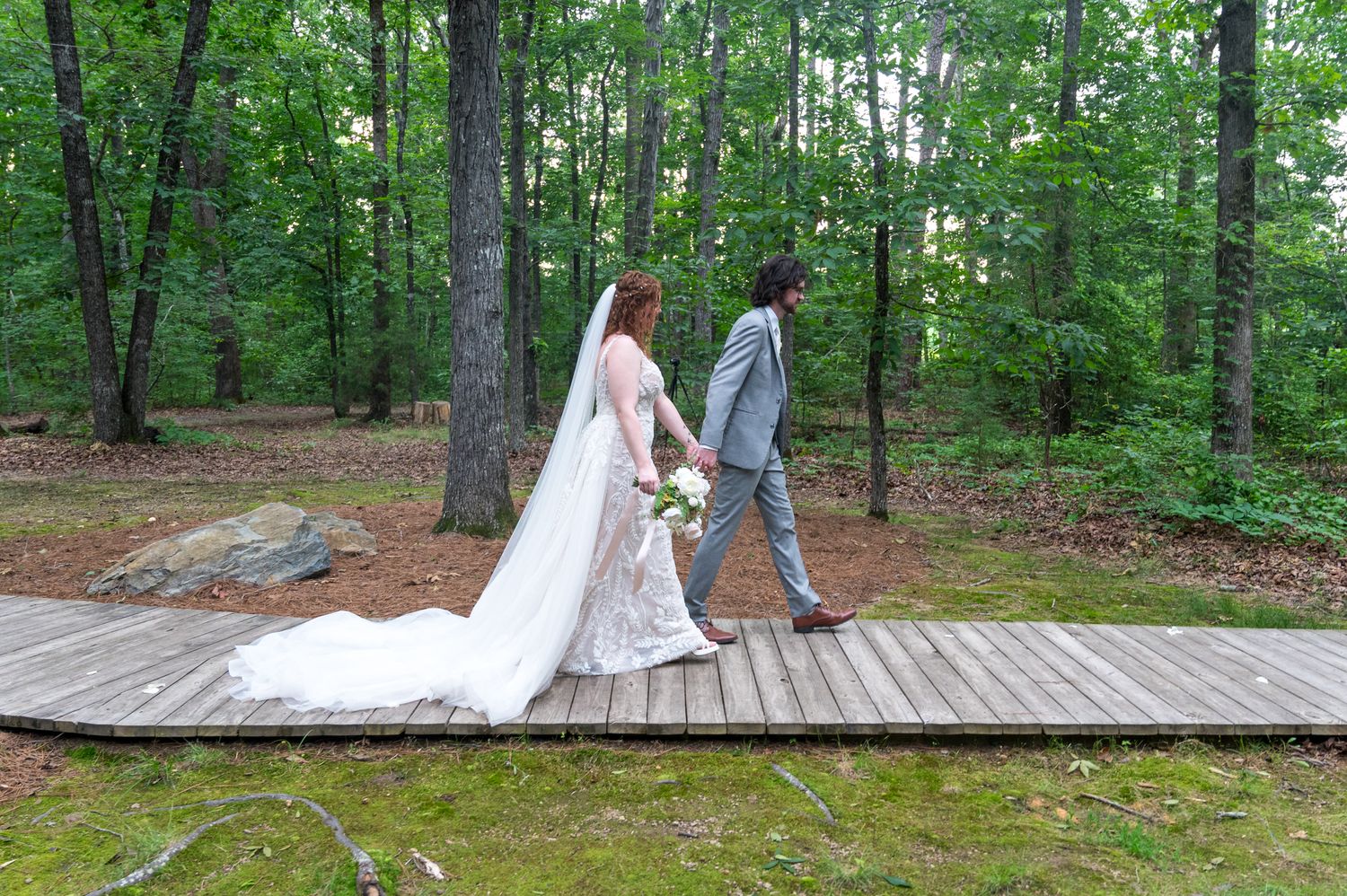 A wedding couple walks along a wooden boardwalk path through a lush forest setting.