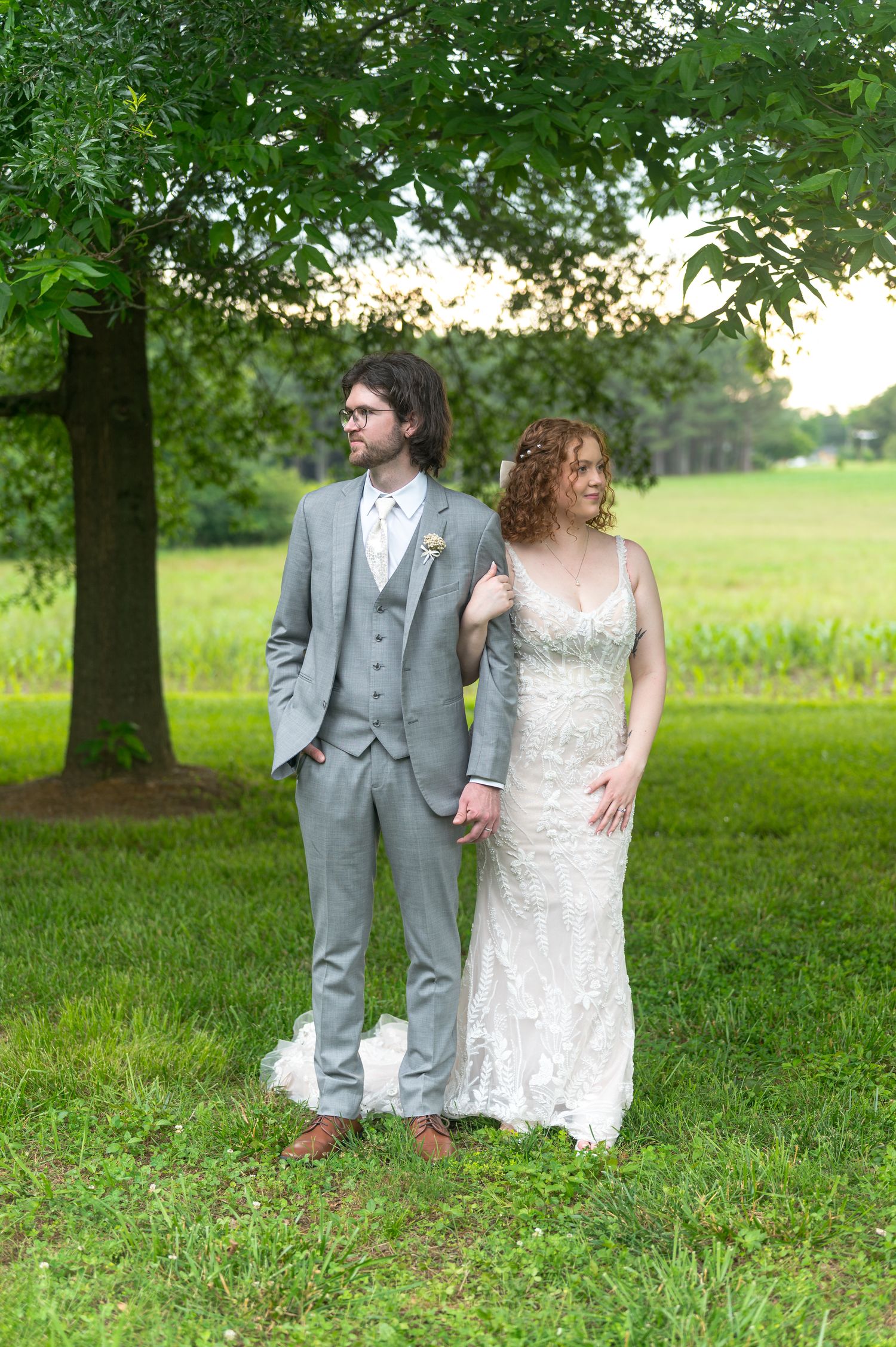Elegant wedding couple poses in grey suit and white dress under tree in pastoral setting.