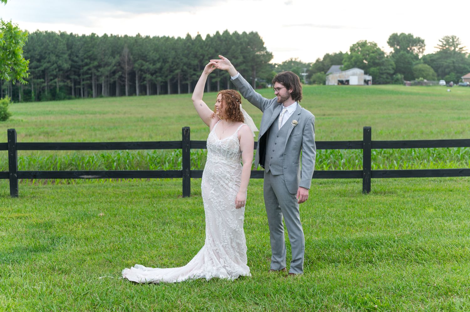 A romantic wedding dance pose on green lawn with rustic fence and forest backdrop.