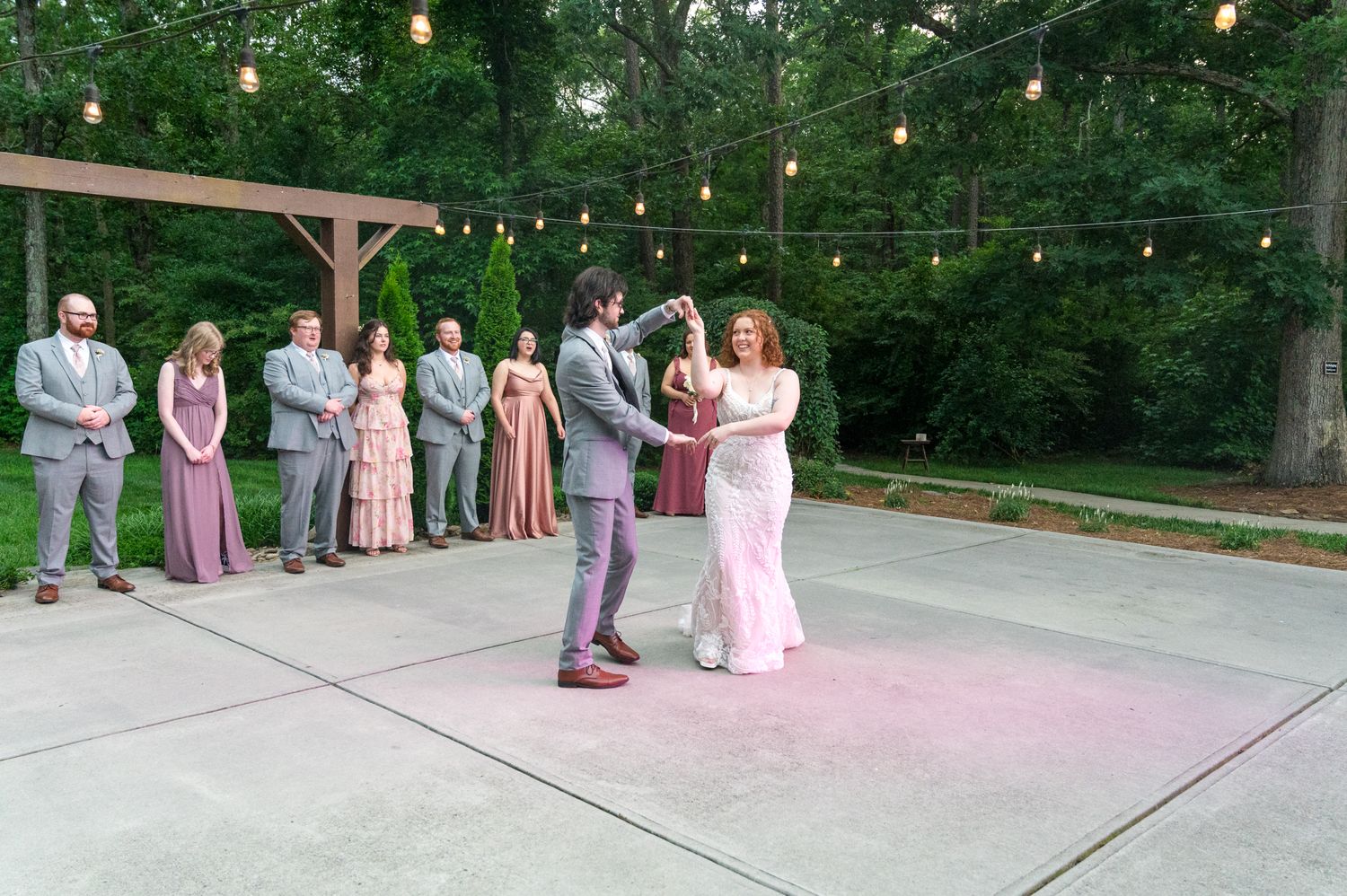 Wedding couple shares first dance on outdoor patio under string lights with wedding party watching.