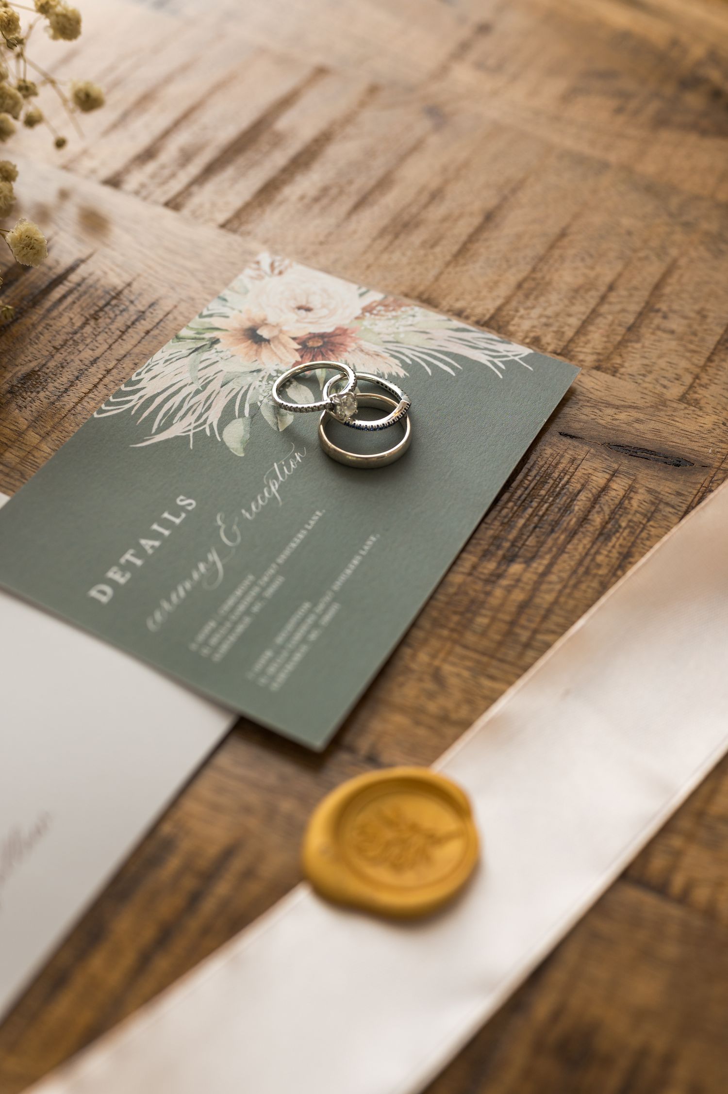 Wedding invitation with dried flowers and wedding rings on wooden surface with gold wax seal.