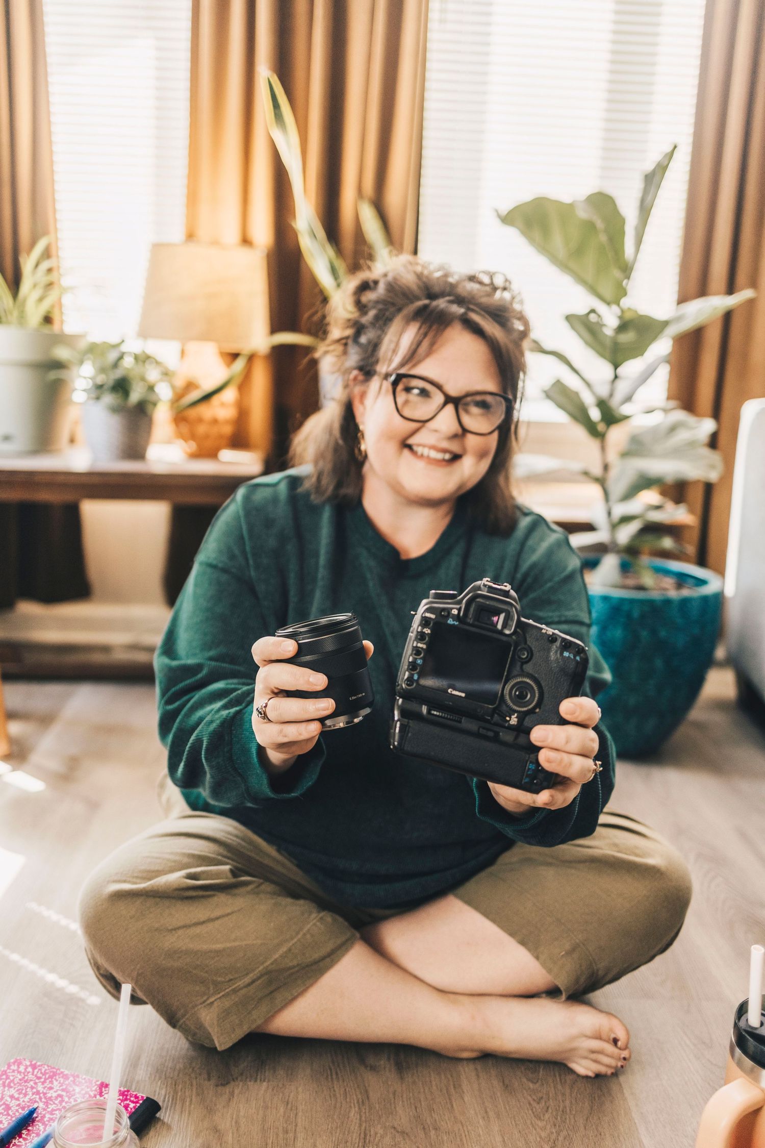 Smiling woman with glasses sitting cross-legged holding DSLR camera and lens.