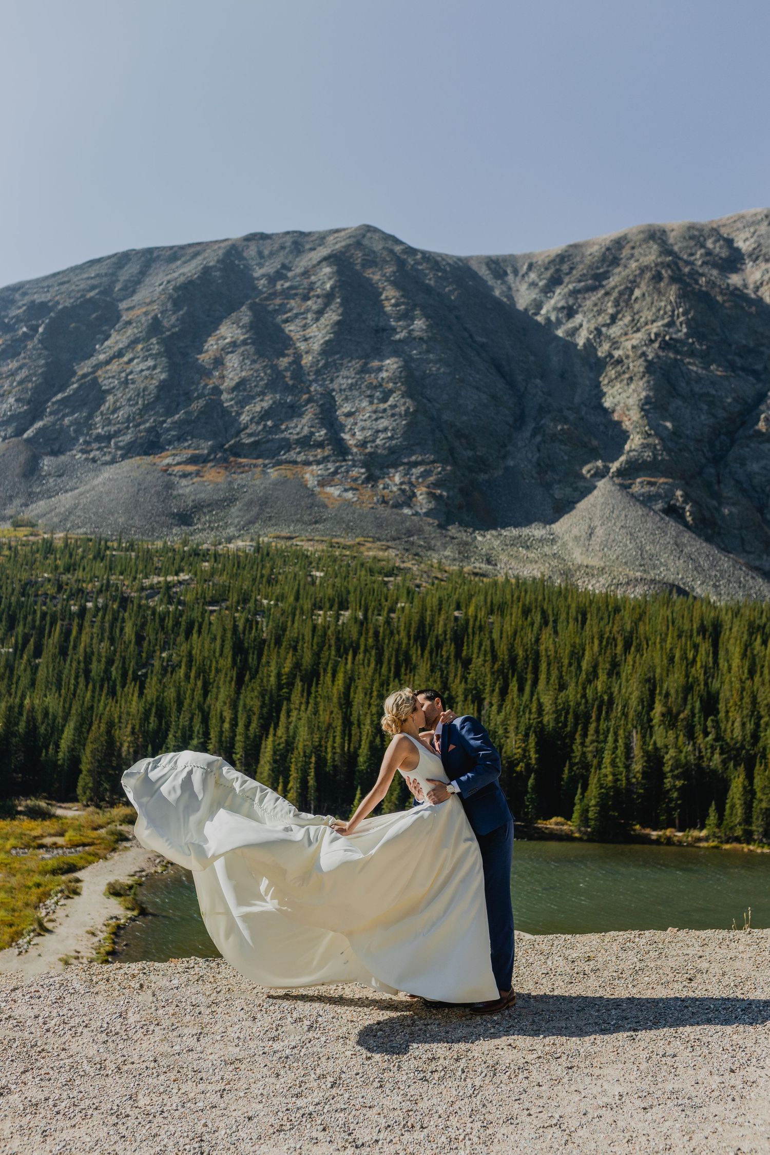 Dramatic mountain wedding photo with flowing white dress against rocky peaks and evergreen forest.