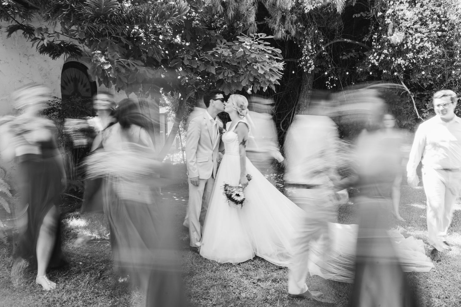 A blurred motion sequence of people dancing and moving in a garden or outdoor setting.