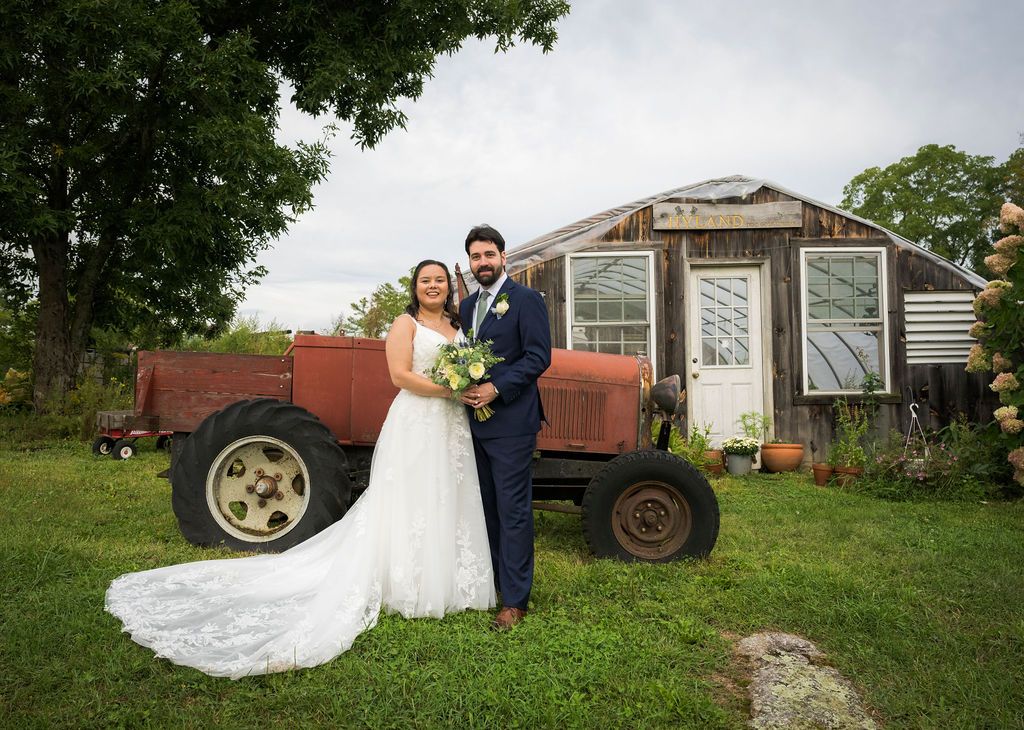 A couple poses in formal wedding attire in front of a rustic red tractor and vintage greenhouse on a grassy lawn.