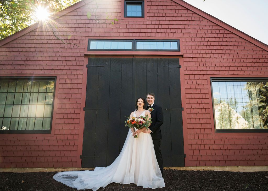 A couple in wedding attire stands in front of a rustic red barn with large black doors and sunlight peeking through.