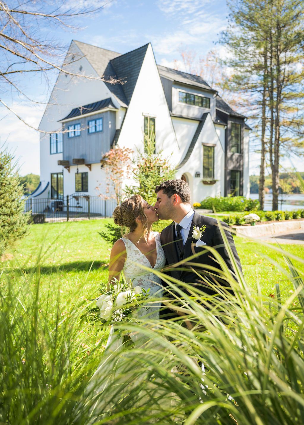 A romantic wedding portrait captured through tall grass with a modern white house in the background.