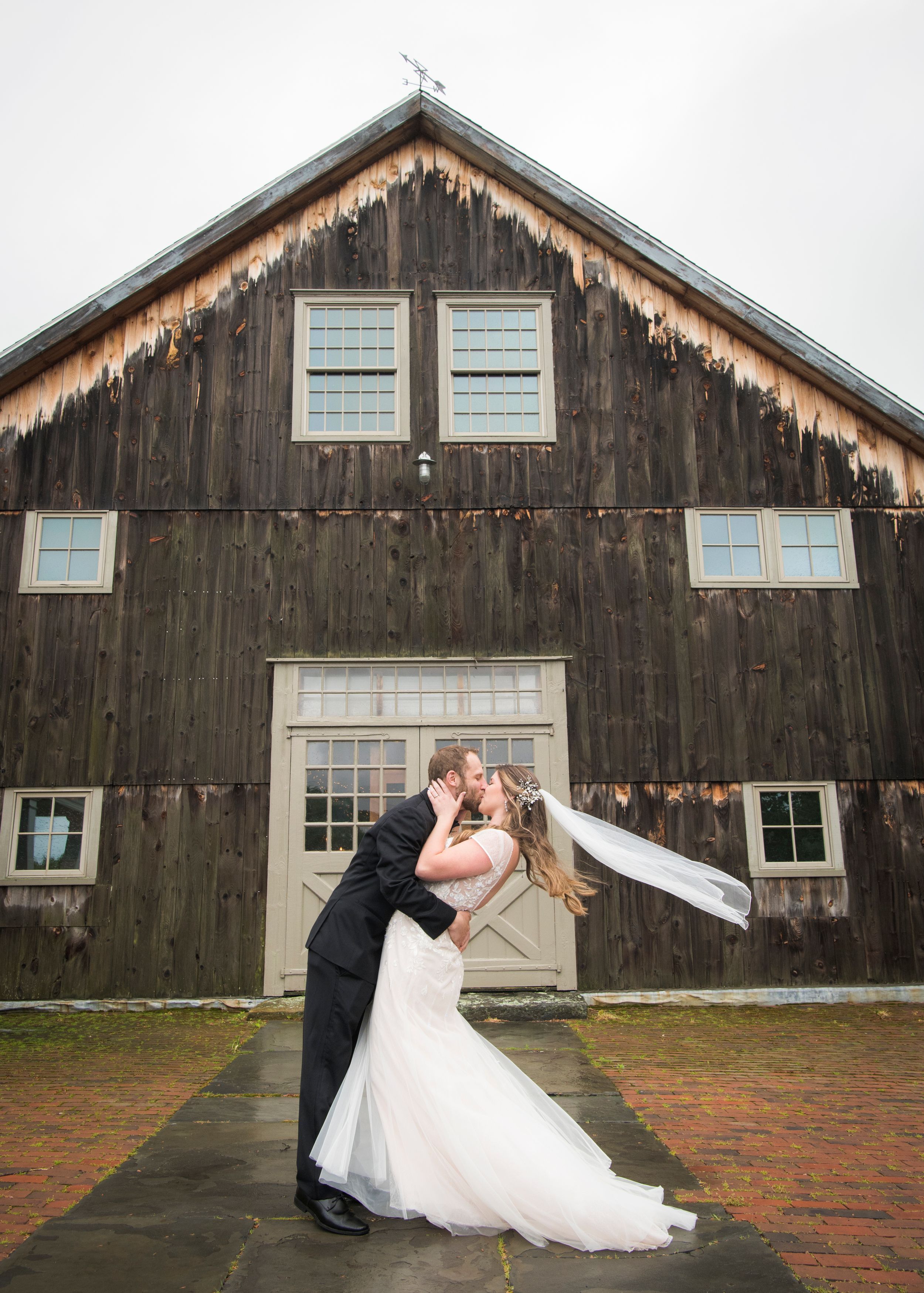 Romantic couple dancing and dipping in front of a rustic wooden barn during a wedding photoshoot.
